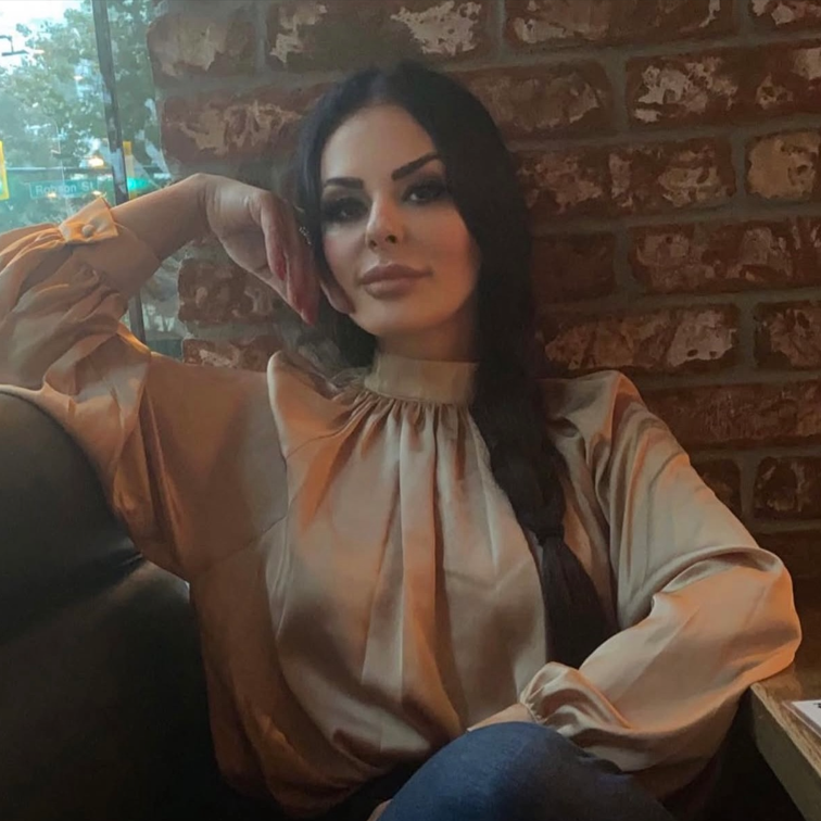 Woman in elegant blouse seated in a cozy café, showcasing a relaxed pose against a brick wall, reflecting a luxurious aesthetic relevant to medical spa branding.