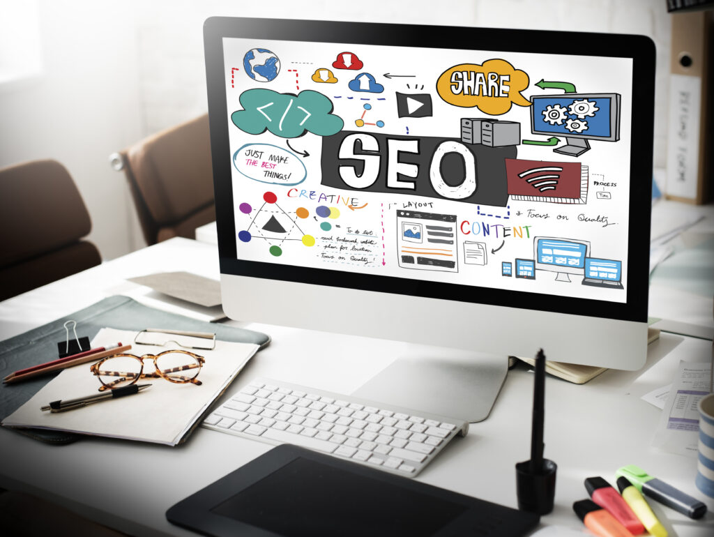 Desktop computer displaying colorful SEO concept graphics, including keywords like "Share," "Content," and "Creative," with a workspace featuring glasses and stationery, emphasizing digital marketing strategies.
